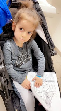 Pompe clinic days | Pompe Disease News | Keara's son, Cayden, sits in a stroller while attending a recent clinic day. He is wearing a gray Nike shirt and holding a coloring page featuring Spiderman.