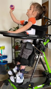A 3-year-old boy with infantile-onset Pompe disease stands upright in a standing adaptive device. The photo is a side view of the boy, who wears a white and orange T-shirt and holds some type of ball in his right hand, as if he's ready to throw it. The stander is a lime green and black color.
