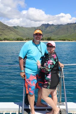 Dwayne and his wife, Jean, pose on the bow of the catamaran. They are both swearing swim clothes, hats, and sunglasses, and in the background is the island of Oahu.