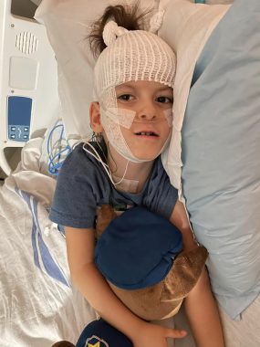 Cayden lies in a hospital bed for a sleep study. He has gauze around his head and wires connected all over his body. He wraps his arms around a large teddy bear