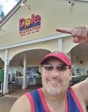 A goateed man with a red visor and blue and red tank top smiles and points to the main entrance of the Dole Plantation in Oahu, Hawaii.