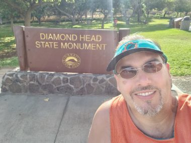 A short brown sign on a shorter stone wall reads "Diamond Head State Monument." In the foreground is a man in sunglasses, a green visor, and an orange tank top. Behind the sign is a green field with trees.