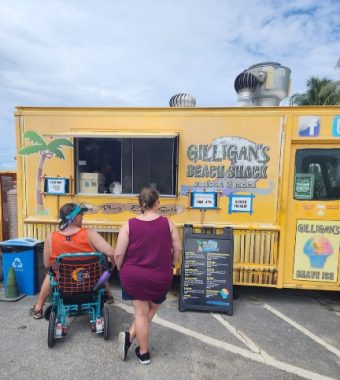 There is a yellow truck with the words "Gilligan's Beach Shack" on its side and a door, with a palm tree on one end. There is a Facebook logo above the door. A man in an orange tank top in a wheelchair is at the left, and a woman in a maroon outfit is on the right; they are facing the serving area. A black menu with white and blue lettering is before them.