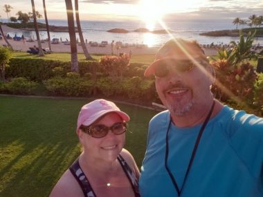 A sunset selfie of a woman and man standing in front of the ocean at a seaside hotel. Palm trees and a sandy beach are in the background. 