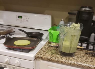 A photo of a kitchen countertop and stove; a blender sits on the counter with some green stuff in it. In a pan on the stove is something that vaguely resembles a pancake.