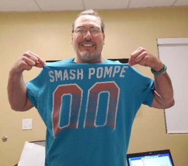 A man smiles and poses for a photo while wearing a blue T-shirt that reads "Smash Pompe"