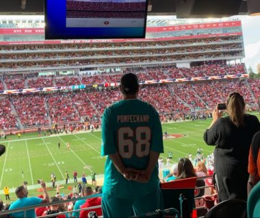 Dwayne stands in front of a railing at Levi's Stadium, overlooking a football game between the Miami Dolphins and the San Francisco 89ers. He's wearing a baseball cap and Dolphins jersey, which has the number 68 and the words "Pompe Champ."