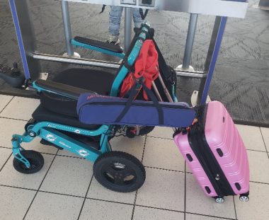 A blue electric wheelchair, loaded with several pieces of luggage, sits near the gate at the airport ahead of boarding.