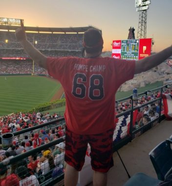 This photo shows a man with his back to the camera and arms outstretched in the stands of a baseball stadium; a scoreboard is visible to one side. He wears an orange T-shirt with blue numbers -- 88 -- and the words "POMPE CHAMP."