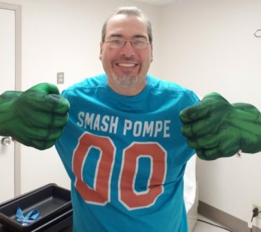 This photo shows a grinning man with brown hair, glasses, and a goatee in a face-and-torso shot. He wears a blue T-shirt with the words "SMASH POMPE" and and orange numbers -- 00 -- with a white border. He wears large hand-and-forearm costume pieces that look like the Hulk's. He's in a room with white walls; a black tray of some sort is behind him to one side.