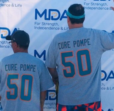 This photo shows the backs of two men, one significantly taller than the other, in front of a banner or wall with several images reading "MDA For Strength, Independence & Life." Each man wears a gray T-shirt with the number 50 and the words 'CURE POMPE," as well as a cap, for the shorter man, and a visor for the other.