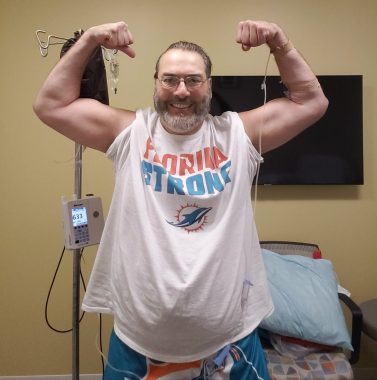 A white man in a Miami Dolphins T-shirt flexes his muscles in the classic bicep pose. He has a beard and glasses, and is smiling broadly. He's standing in a clinic infusion room, next to a chair with a pillow, and has an IV line connected to an IV pole.