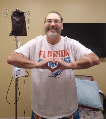 A white man with a beard and glasses and a Miami Dolphins T-shirt stands in front of an infusion chair and IV pole in the clinic. He makes the shape of a heart with his two hands in front of his chest. 