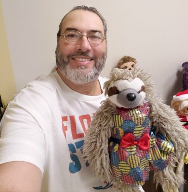 A white man with a beard and glasses and wearing a Miami Dolphins T-shirt takes a selfie with a stuffed animal sloth on his lap. 