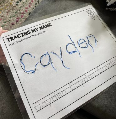 The photo shows a white piece of paper with the word "Cayden" at the center, with a marker tracing over it. The words "TRACNG MY NAME" are at the top left, with "I can trace and write my name" beneath it. A cartoon pencil drawing is at the top right. At the bottom are three well-written "Cayden"s within two lines, with another line in the middle to mark the height of the little letters. 