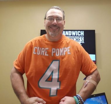 A photo shows the face and torso of a man with light brown hair, glasses, and a goatee. He wears an orange T-shirt with the number 4 and the words "CURE POMPE," as well as several strings of beads and another bracelet on one wrist. He appears to have an IV syringe taped onto one forearm. Behind him is a tannish wall with a black sign that can be partly read; it apparently says "Sandwiches."