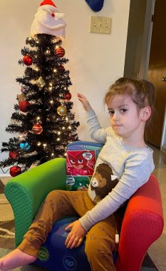 A young boy with a cute buffalo pajama shirt sits in a small red and green chair in front of a small decorated Christmas tree. 