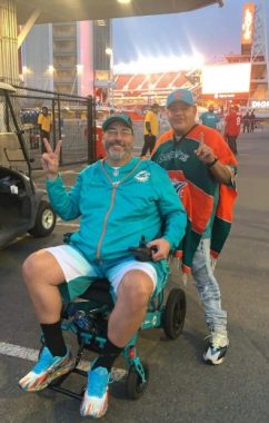 Two men smile for a photo in front of Levi's Stadium after watching a Miami Dolphins football game. Dwayne is wearing a turquoise jacket and sitting in his electric wheelchair. His friend, Louie, is standing next to him wearing a striped poncho and baseball cap.