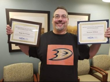 A man with short hair, glasses, and a goatee stands in an office holding two framed certificates. He's smiling and wearing a black sports T-shirt with an orange and gold design at the center. Chairs and a couple of pictures are against the wall behind him.