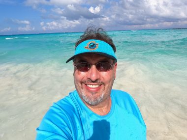 A selfie shows a man's head, shoulders, and chest as he stands in clear and turquoise water. He's wearing a teal shirt and visor and tinted sunglasses. While some clouds are gathering, it appears to be a sunny, warm day.