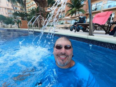 In this photo, a man's head has emerged from the water in a pool, with buildings, pool chairs, and a wheelchair in the background. He's wearing a blue shirt and tinted sunglasses. A small waterfall is splashing just next to him.