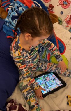 A young boy in pajamas sits on a bed or a blanket and looks down at a game on a tablet. His brown hair is long and braided. 