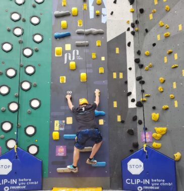 A photo shows a man in a black T-shirt, navy blue shorts, tennis shoes, and a yellow helmet climbing a gray artificial wall with yellow, blue, and gray footrests. He appears to be near the bottom. On each side of him are other climbing walls.