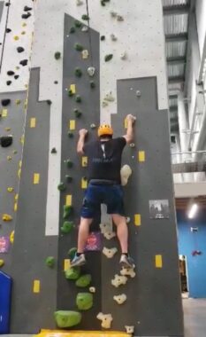 This photo shows the man on his journey up a wall, this one with white areas near the top and some green and white footrests. To his right we see the high ceiling of the indoor facility, with a blue wall and doorway in the distance.