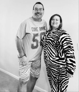 A black-and-white photo shows a white, late middle-aged couple standing side by side in front of a white wall. The man, wearing a goatee, glasses, and a T-shirt that says "Cure Pompe" and "50," smiles while standing next to the woman, who also is smiling and wearing a zebra-print outfit.
