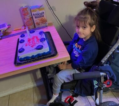 A five-year-old boy sits in an assistive device in front of a table with a white birthday cake with blue frosting balloons. He glances up and to his left at the camera. 