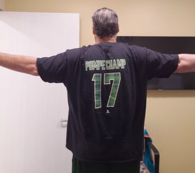 A man stands with his back to the camera and holds his arms straight out to the sides. He's wearing a black T-shirt that reads "Pompe Champ 17" on the back in green lettering.