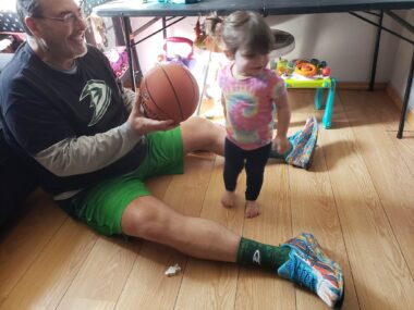 A man sits on a wooden living room floor and leans against a couch. His legs are spread in a V-shape, and he's holding out a basketball to a toddler, who's standing between his legs. 