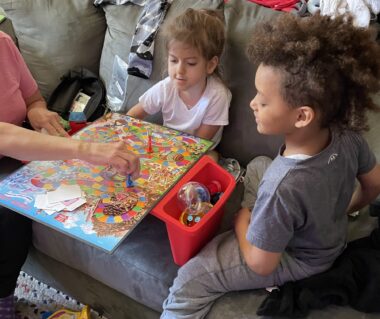 Two young boys sit on a couch playing Candy Land with a woman, seen by her arms and part of her chest. The larger boy on the right is dressed in gray; the smaller boy at center wears a white shirt.