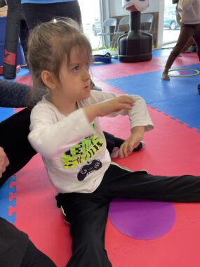 A 5-year-old boy sits on a pink, purple, and blue gym mat, legs spread, and arms extended in a karate pose. He looks intently up and to the left, presumably at the karate instructor, as it's a beginners' karate class.