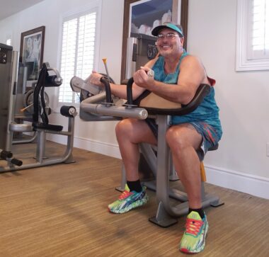 A guy in the gym smiles at the camera while sitting at a bicep curling weight machine. He's wearing glasses, a visor, a tank top, shorts, and athletic shoes. The clothing color scheme is of the Miami Dolphins. 