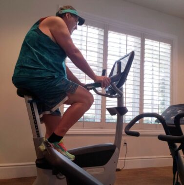 A side view of a man in a gym riding a stationary bicycle. He's wearing shorts and a tank-top and visor with the colors of the Miami Dolphins. 