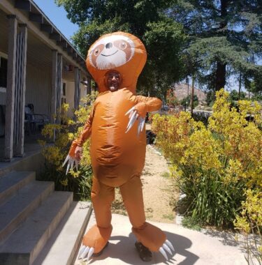 A man wears a full-body, inflatable, orange sloth costume while posing in a garden outside a house. 