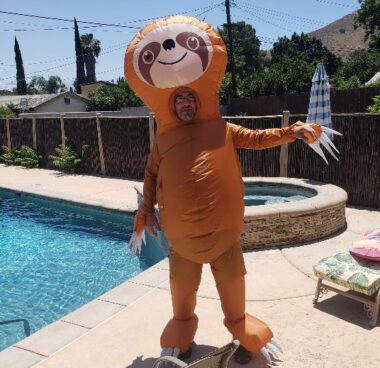 A man wears a full-body, inflatable, orange sloth costume while standing next to a pool.