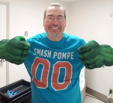 A man with late-onset Pompe disease smiles while wearing a blue T-shirt that says "Smash Pompe." He's wearing Hulk gloves and holding his arms up in a fighting stance.
