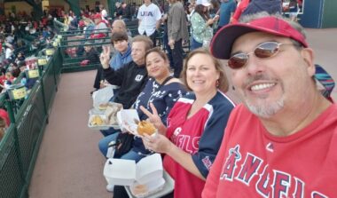A group of five people smile for a selfie while sitting in a stadium at a baseball game. There are two couples, Dwayne and Jean, and Scott and Maritoni, as well as Scott and Maritoni's son, Peyton. Dwayne and Jean are wearing red Angels shirts, and all appear to have styrofoam containers of food in their laps.