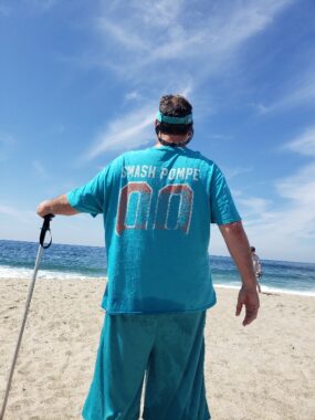A man stands on a beach with his back to the camera. He's wearing a blue shirt that says "Smash Pompe" on the back, and he's leaning on a ski pole with his left arm. It's a bright, sunny day, and he's looking out toward the ocean.