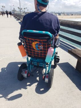 A man with his back turned to the camera sits in a blue wheelchair on a pier, looking toward the beach.