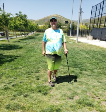 A man wearing a green and blue tie-dye T-shirt and green shorts walks across the grass at a park while using his hiking stick for support. There appears to be a baseball field to his left, and gentle hills rise in the background.