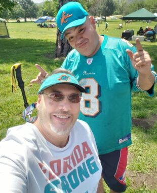 Two men pose for a selfie in the park while attending a Miami Dolphins fan club barbecue. Dwayne is seated in his wheelchair while his friend Louie stands beside him. Both men are decked out in Dolphins apparel.