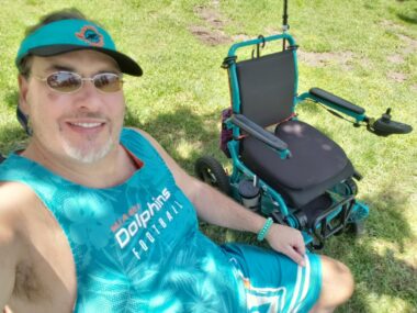 A man decked out in aqua Miami Dolphins apparel sits in the shade at a park. His wheelchair, also aqua to represent the Dolphins, is parked beside him.