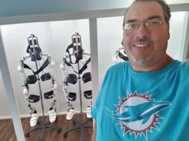 Two skeletal, white and black robots are at the back of a room, in one part of a white storage area. In the right foreground is a man with short brown hair, glasses, and a teal Miami Dolphins T-shirt.