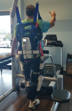 A man strapped in a harness and exoskeleton is seen from the back as he walks.