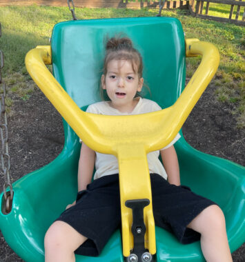 A 5-year-old boy sits in an accessible swing at a local park. The swing is green with a yellow safety bar that extends in front of the boy and over his shoulders.