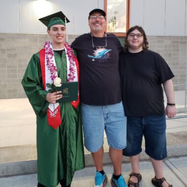 An older man stands between two young men with his arms around them. The one on the left is wearing a green cap and gown and holding a diploma to celebrate his high school graduation. He also has red and white sashes and a lei around his neck. The young man on the right is wearing a black T-shirt and jean shorts, as is the older man in the center.