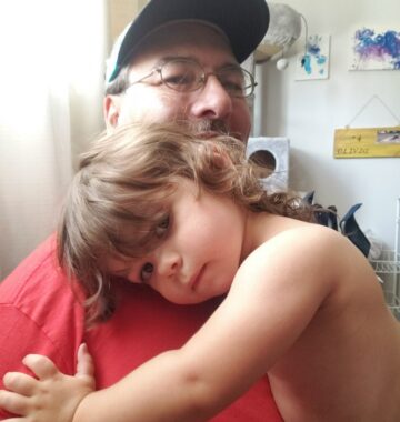 A man snuggles with a 2-year-old. He's wearing a red T-shirt, baseball cap, and glasses, and the girl, without a shirt, has her head resting on his shoulder.